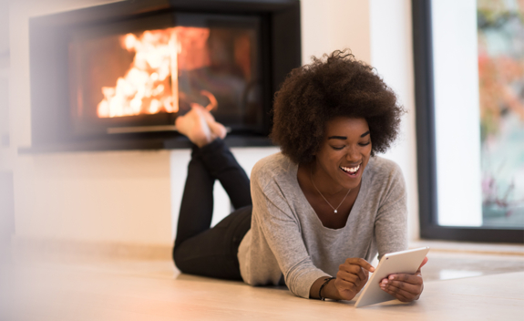 Woman laughing and using a tablet device while laying on floor in living room with fireplace in background
