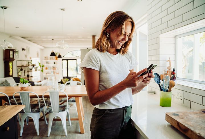 A smiling woman standing in a bright and airy kitchen while using her cell phone